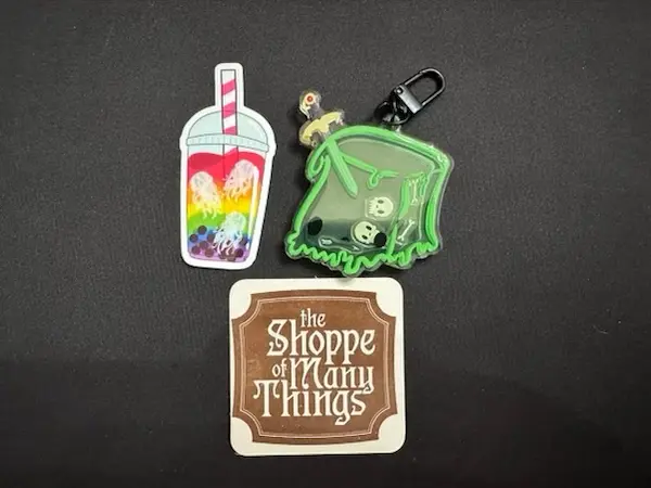 Sticker of cup with straw and floating tentacled creatures. Keychain of green gelatinous cube with sword sticking in it and skulls and bones inside. Logo reading the Shoppe of Many Things.